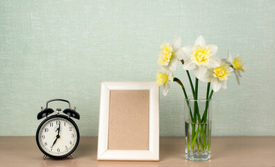 Daffodils flowers in a vase, vertical frame for mocap photos, a mechanical alarm clock on a table against a turquoise wall
