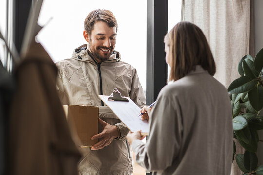 shipping, transportation and people concept - delivery man with parcel box and customer signing papers on clipboard at home