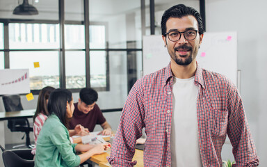 Closeup on caucasian beard handsome creative and business man wearing casual shirt and eyeglasses, crossing arms and smiling with blur background of colleagues working in indoor office or workplace.