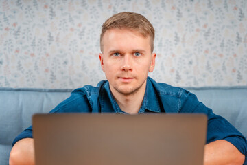 A young man is sitting at a table and looking out from under a laptop. Work on a laptop remotely from home or from a cafe. Technologies, remote work and social networks