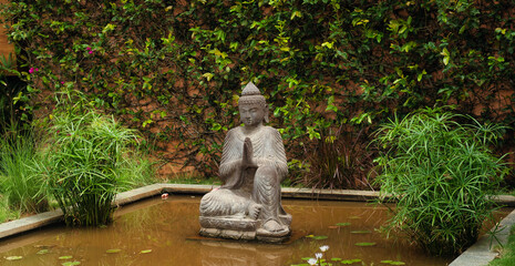 A statue of a meditating Lord Buddha in a garden set amidst water and vegetation