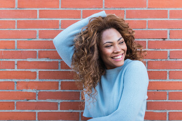 Happy mixed race woman with white perfect smile, long curly hair, glamour makeup and blue jumper posing in brick wall. Beauty woman Latin Ethnic female face. Copy space for text