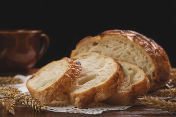 Sliced freshly baked round bread with ears of wheat on white napkin on wooden table on black background, selective focus.