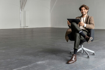 Smiling businessman with digital tablet sitting on chair in empty industrial hall