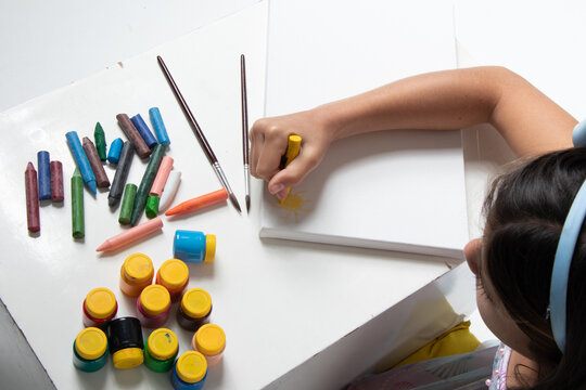 cute girl with light blue bow on her head, painting a blank canvas with yellow crayons, drawing the sun, photo view from above.