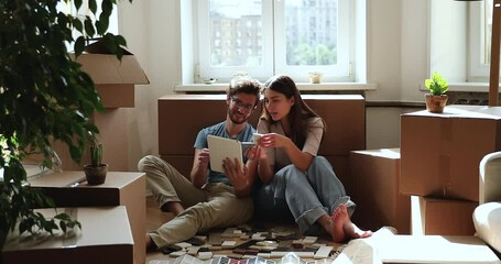 Inspired married couple do renovation at own apartment sit on floor among packed belongings choose materials online use tile samples paper web catalogues on pad. Excited homeowners redecorate new flat