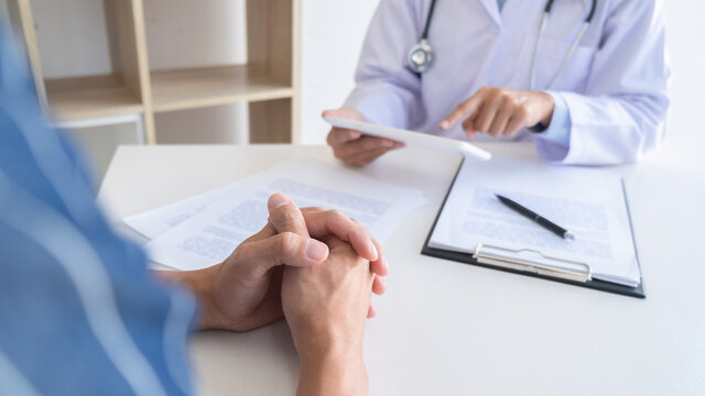 Patient Listening Intently To A Male Doctor Explaining Patient Symptoms Or Asking A Question As They Discuss Paperwork Together In A Consultation