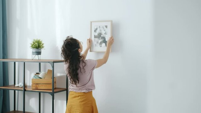 Slow motion of creative lady hanging picture on wall in new modern apartment after relocation and enjoying art. People and housing decoration concept.
