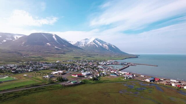 Wide aerial shot of a coastal town in Iceland. Turns to reveal bright sun and mountains. Beautiful clear day.