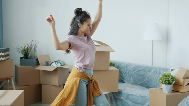 Slow motion portrait of excited young woman dancing in new apartment after relocation with cardboard boxes and packed furniture in background.