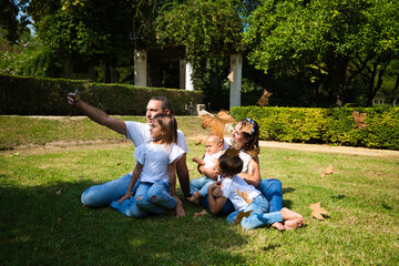 Obraz premium family portrait of five members, father, mother and children sitting on the grass taking a photo with the mobile phone while the leaves fall from the trees in autumn. Family concept.