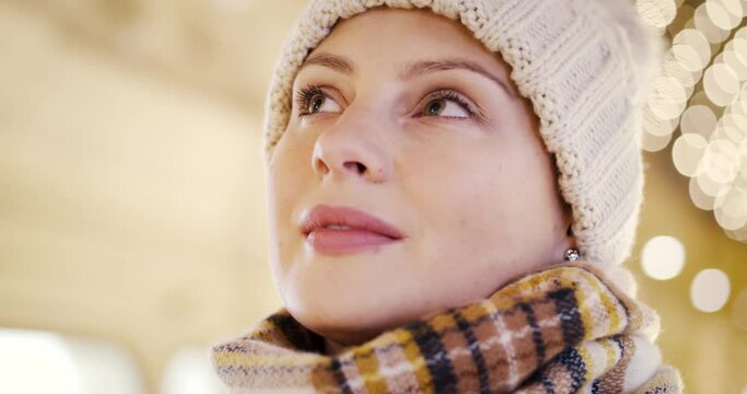 Close up divinely inspired face of young girl in hat and scarf thinking about dreams. Christmas New Year theme background with festive holiday bokeh lights. People hopes, makes a wish