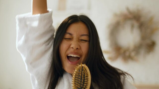 Webcam pov portrait of young playful asian woman singing to camera with hairbrush, pretending to be popstar at home