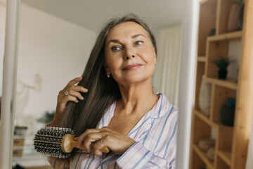 Beautiful, charming elderly female combing grey hair with round hairbrush standing against shelves in hallway or bedroom in front of mirror, looking at her reflection, getting ready to go to bed - 472072971