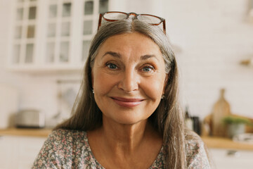 Webcam picture of charming stylish natty grandmother with nude daily make-up, looking at camera with lovely, warm smile, sitting against kitchen interior background, talking to her friend