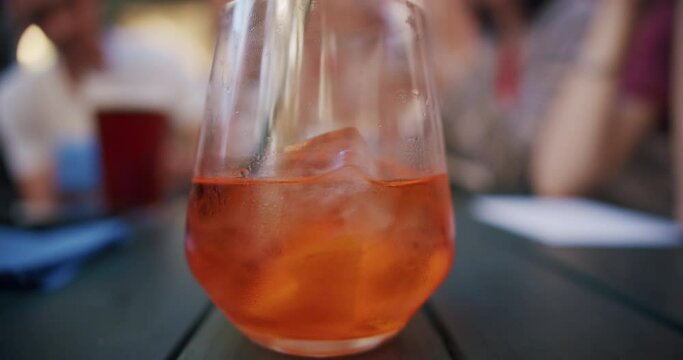 Close up of mist up cocktail glass with an orange aperol spritz with ice on the table in a summer cafe. The hand stirs the drink with a straw. Slow motion. Leisure time in cafe and bar
