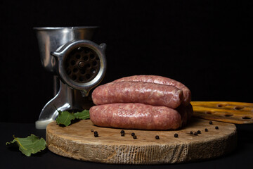 Raw meat sausages on a wooden board on a black background. Homemade sausages in a natural casing next to a steel meat grinder. Meat products. Home cooking