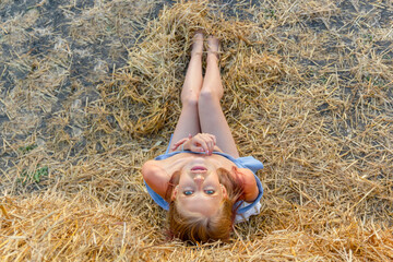 A young beautiful blonde girl with blue eyes and snow-white skin sits on the hay on the ground and her head thrown back looks relaxed up. Summer sunny day