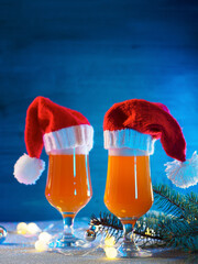 Two glasses of Christmas beer ale in Santa hats on the festive table. Blue wooden background, garland and branches of the Christmas tree. Copy space