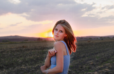 Fototapeta na wymiar A young beautiful blonde girl with blue eyes and snow-white skin sits on the ground, leaning on a haystack, looks relaxed to the side enjoying the rays of the setting sun in summer.