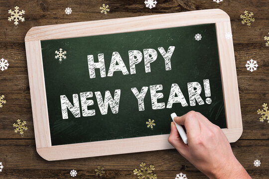 hand is writing message HAPPY NEW YEAR on a chalkboard surrounded by decorative snowflakes on wooden background