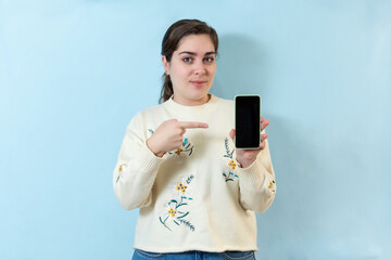 Smiling positive young woman shows a blank screen smart phone. Isolated on light blue background.