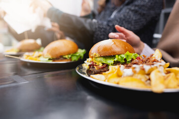 Close-up side view burgers menu with french fries on table. Unrecognizable people on background with light flare.