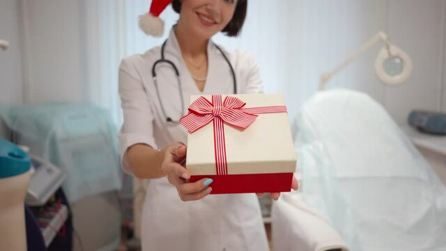 dermatologist doctor in lab coat and Santa Claus hat holds Christmas gift in her hands in medical office. New Year 2022. Advertising of medical clinic, gifts and discounts for new year. Christmas