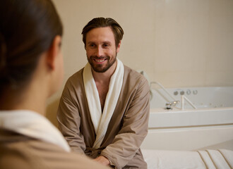 Handsome middle aged European man in bathrobe smiling looking at his wife, out of focus on the foreground, resting at wellness spa resort during honeymoon. Couple getting beauty treatment together