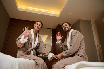 Beautiful happy middle aged heterosexual couple in love, in bathrobes, holding hands, sitting on massage tables and waving, smiling, looking at the camera. Newlyweds honeymoon at spa resort