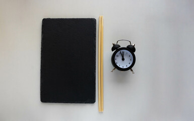 A black slate plate with chopsticks and a black alarm clock. A plate of sushi on a white table. Place for text