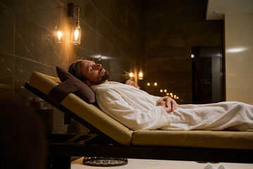 Attractive handsome middle aged man in bathrobe relaxing with closed eyes, lying on a chaise lounge near a blurred woman on the background, in a luxury serene atmosphere of a wellness spa center