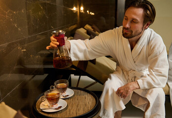 Relaxed handsome bearded middle aged Caucasian man relaxing in spa lounge sitting in white terry bathrobe on sun lounger and pouring herbal tea into glasses from transparent glass decanter