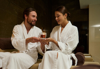 Handsome loving man giving a cup of herbal tea to her beloved wife, resting together in lounge area by jacuzzi and swimming pool in luxury wellness spa resort, enjoying closeness on their honeymoon