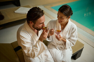 Beautiful loving couple of European man and woman, in white bathrobes, chilling, sitting close to each other by jacuzzi in luxurious wellness spa resort, quietly talking over cup of healthy tea drink
