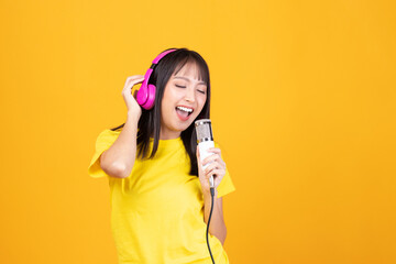 Pretty Asian woman wearing yellow shirt holding microphone singing karaoke music on yellow background. Listen music with headphones sing song in microphone.