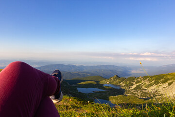 Naklejka premium Against the backdrop of green mountains and mountain lakes and a view of the valley above the clouds, the legs of a human are located in the foreground. Active rest climbing mountains on foot.