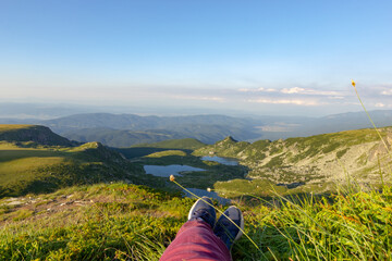Naklejka na ściany i meble Against the backdrop of green mountains and mountain lakes and a view of the valley above the clouds, the legs of a human are located in the foreground. Active rest climbing mountains on foot.
