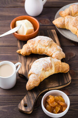 Fresh crispy croissants on a wooden board, a cup of coffee and jam in a bowl on the table. Brunch and coffee break. Vertical view
