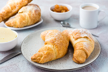 A pair of fresh crispy croissants on a plate, a cup of coffee and bowls of jam and butter on a light textured background. Coffee break or breakfast