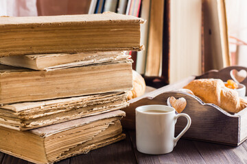 A large stack of old books, a cup of coffee and a tray of croissants on a wooden table. Relaxation, wellness and home reading.