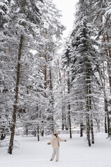 Happy girl in warm clothes spinning around on snowy ground in winter forest. Trees covered by snow. Happiness concept