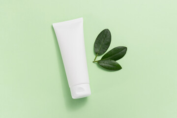 Mockup white tube of face cream with green leaves top view, light green delicate background