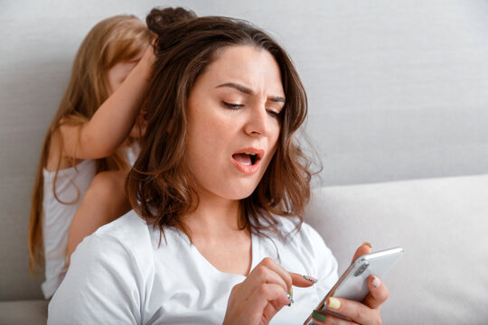 Young mother with internet addiction using smartphone while unhappy stressed daughter pulls her hair