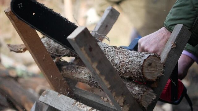 A footage of a hand holding a saw cutting beech firewood. Preparation of firewood in the yard. Cutting beech logs with a chainsaw. In the background another man is chopping firewood with an axe