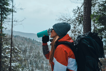 Rest in the mountains. Beautiful young woman dressed in warm sportswear, hat and sunglasses drinks...