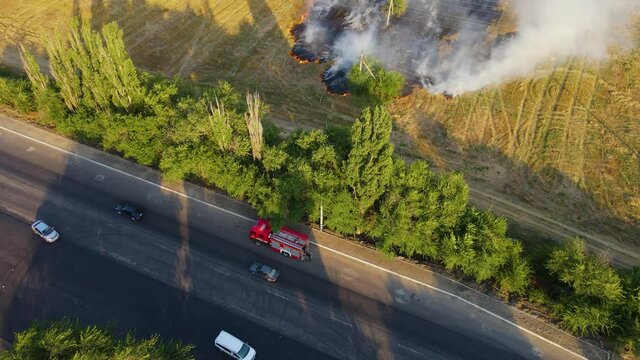 Cinematic aerial view firefighters pull fire hoses to extinguish huge burning flame in field with dry stubble near highway with moving cars. Emergency case for danger mission and rescue nature saving