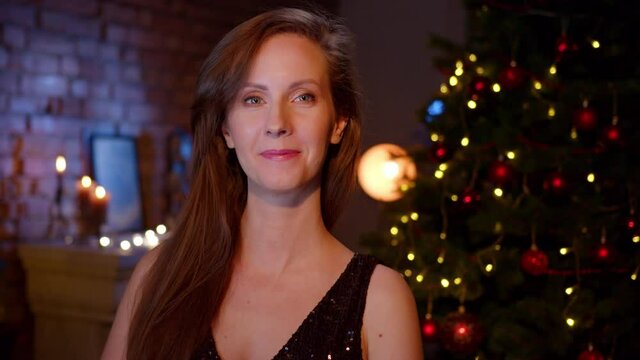 Happy woman wishing Merry Christmas and Happy New Year. Portrait of woman in elegant black dress at home with Christmas tree.