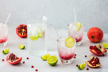 Refreshing pomegranate drink in glasses with pomegranate seeds and ice, lime and glass straws, jug of cold water with citrus, cut pomegranates with seeds