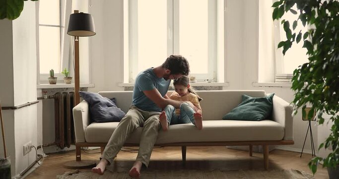 Bonding young foster father playing active game tickling on couch with little adopted child girl. Joyful millennial dad having fun with small daughter laughing messing enjoying funny playtime at home